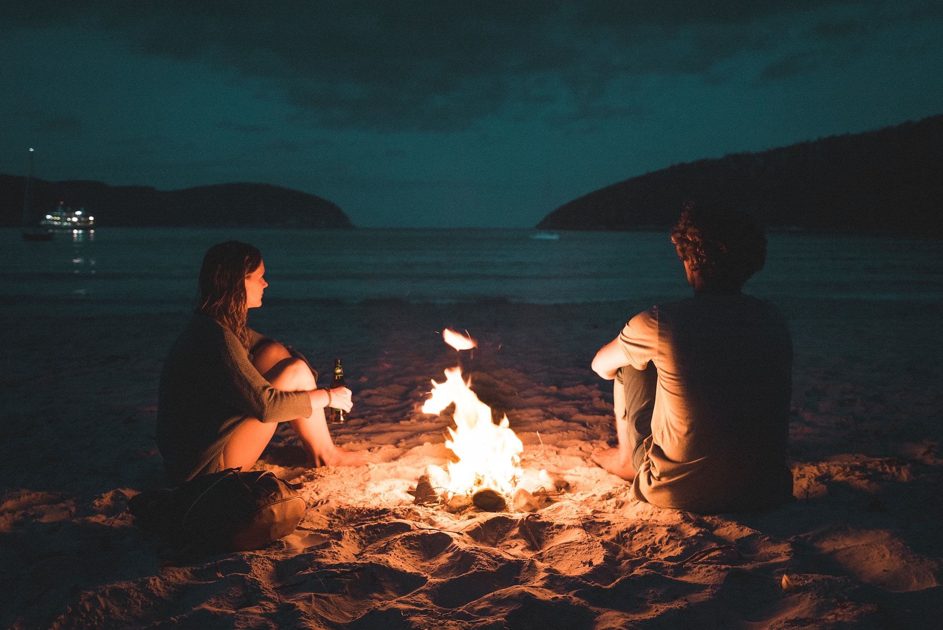Una pareja disfruta de una fogata en la playa. Están sentaos sobre la arena mirando el mar.