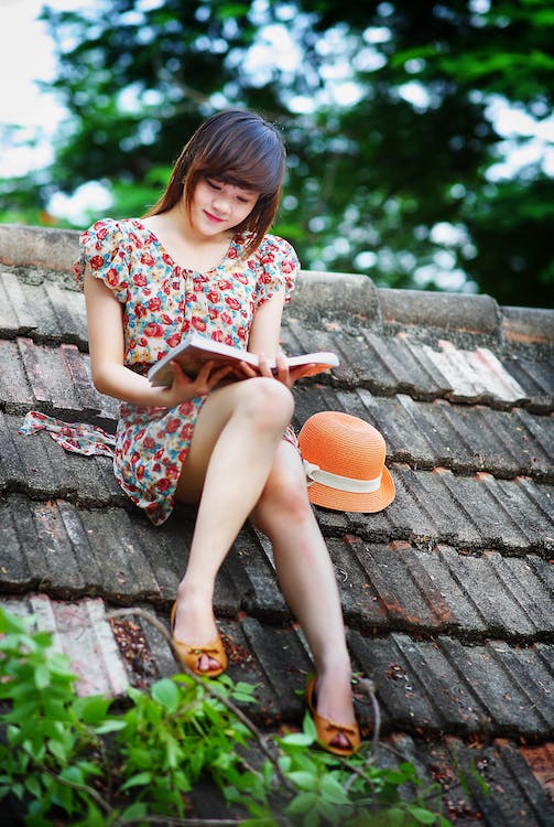 Una joven sentada en el techo de una casa, lee un libro y viste un traje de flores estampadas.