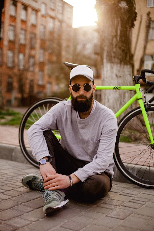 Un joven sentado en la calla, Una bicicleta al fondo. Lleva gafas y viste ropa urbana.